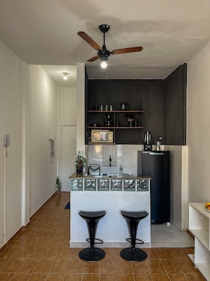 A compact kitchen with a ceiling fan, a coffee maker, and a microwave. There are two stools at the counter. - Studio vista mar pertinho da praia (Rio de Janeiro)