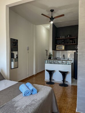 A bedroom with a bed, a ceiling fan, a bathroom with a towel, and a kitchenette with a microwave and bar stools. - Studio vista mar pertinho da praia (Rio de Janeiro)