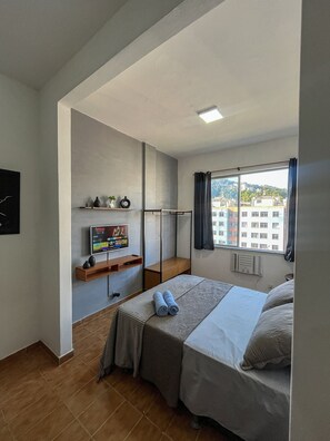Habitación de hotel con cama, televisor, ventana y vistas a edificios, además de un estante con objetos decorativos.