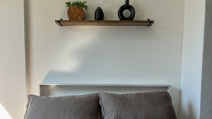 Sängyssä on kuvioitu peitto, kaksi tyynyä, pieni hylly, jossa on kasvi ja kaksi mustaa esinettä, sekä sängylle aseteltuja rullattuja pyyhkeitä.