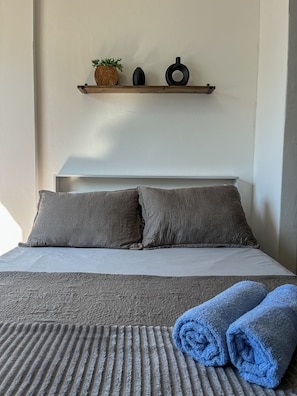 A bed with a textured blanket, two pillows, a small shelf with a plant and two black objects, and rolled towels on the bed. - Studio vista mar pertinho da praia (Rio de Janeiro)