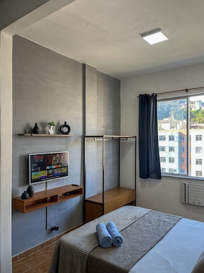 A hotel room with a bed, a TV on a shelf, a window with curtains, and a view of buildings outside. - Studio vista mar pertinho da praia (Rio de Janeiro)