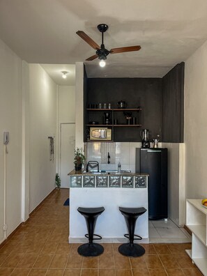A compact kitchen with a ceiling fan, a coffee maker, and a microwave. There are two stools at the counter. - Studio vista mar pertinho da praia (Rio de Janeiro)