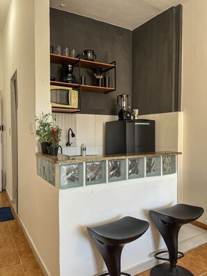 A kitchen with a black refrigerator, a coffee maker, and a blender on the countertop. There are two black bar stools and a potted plant on the counter. - Studio vista mar pertinho da praia (Rio de Janeiro)
