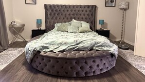 A round, tufted bed with a grey headboard in a bedroom with two bedside tables, a floor lamp, and artwork on the walls.