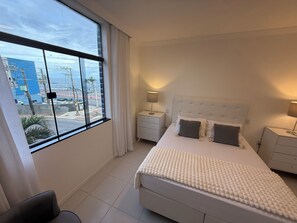Un dormitorio con un ventanal grande, una cama con cabecera blanca, dos mesitas de noche y vistas a la playa y edificios.