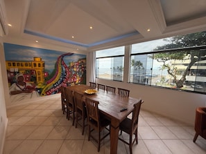 Un comedor con una mesa grande y sillas, un mural en la pared, y vistas a la playa a través de amplios ventanales.