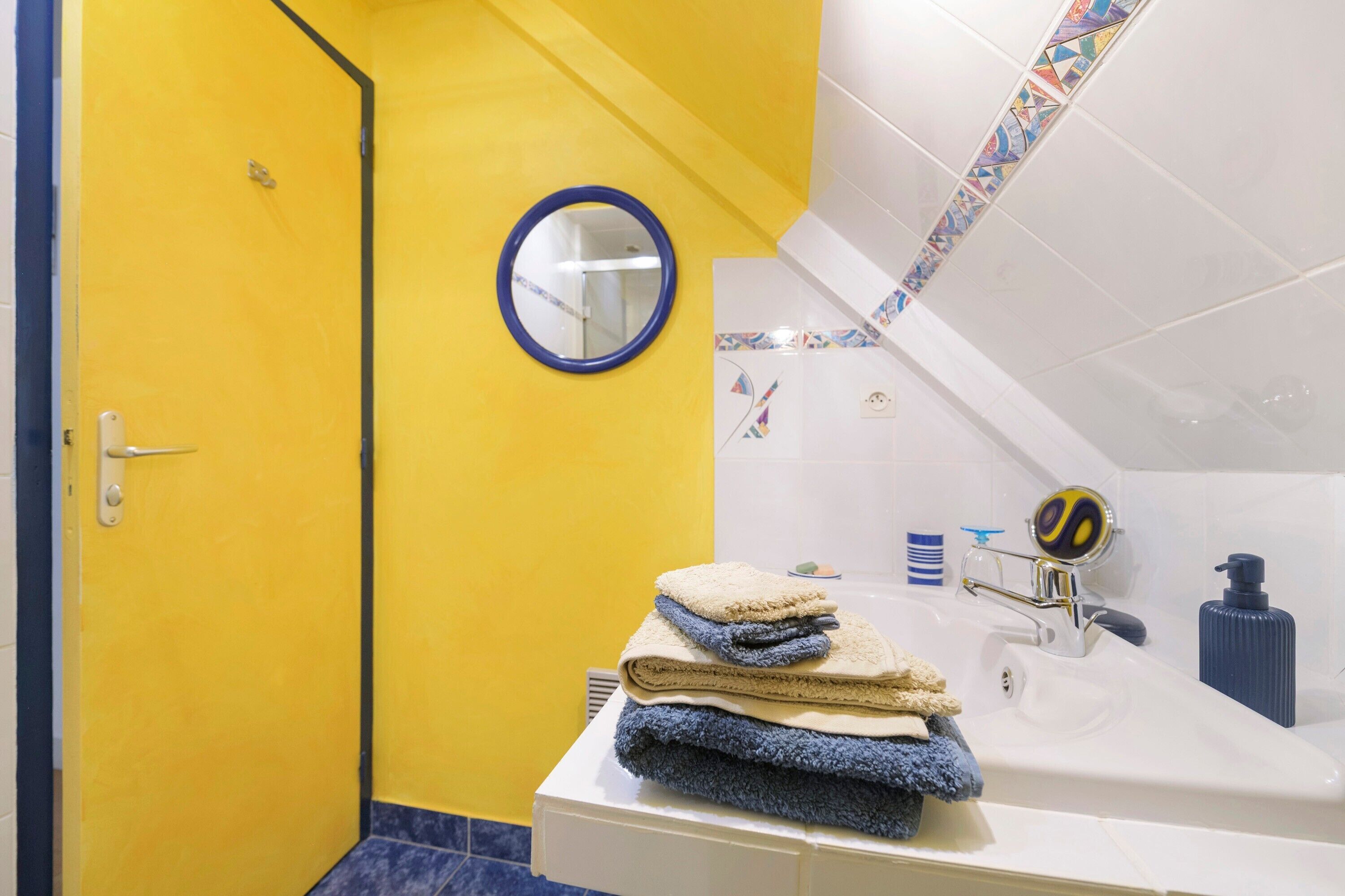 Une salle de bain avec une porte jaune, un lavabo blanc avec un robinet bleu, un miroir et une pile de serviettes.