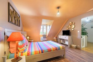 Une chambre avec un plafond en pente, un lit recouvert d’une couette à rayures colorées, une télévision, un bureau avec une lampe et une étagère fixée au mur.