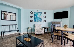 A living room with a black coffee table, a floral armchair, a dining table with chairs, a TV, and a wall-mounted mirror. - Oceanfront Suite w/Balcony-Resort Pools&Lazy River (Myrtle Beach)