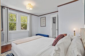 Ein Schlafzimmer mit einem großen Bett, einem Fenster mit Vorhängen, einem blauen Hocker, einem gerahmten Zertifikat an der Wand und einer Nachttischlampe.