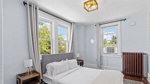 Quarto com cama, mesas de cabeceira, radiador e janelas grandes.