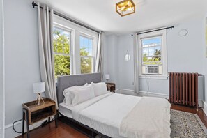 Ein Schlafzimmer mit Bett, Nachttischen, einem Heizkörper und großen Fenstern.