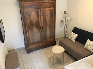 A room with a wooden wardrobe, a sofa, a small stool, and a floor lamp. - Cottage "Little House in Coulon, Marais Poitevin" (Coulon)