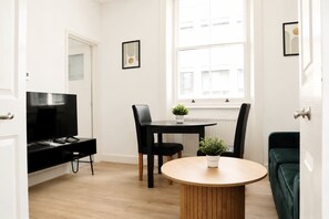 A modern living room with a TV, a dining table, chairs, a small plant, and a green sofa. - Bright 1BR in Fitzrovia, Steps from Oxford Street (London)