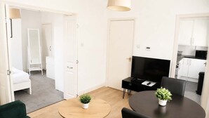 Ein modernes Wohnzimmer mit Fernseher, zwei Topfpflanzen, einem runden Holztisch, einem grünen Sofa und einer Küche im Hintergrund.