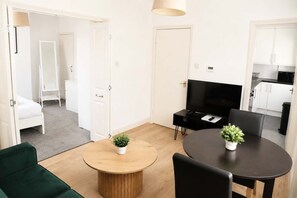 Ein modernes Wohnzimmer mit Fernseher, zwei Topfpflanzen, einem runden Holztisch, einem grünen Sofa und einer Küche im Hintergrund.