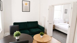 Ein Zimmer mit einem grünen Sofa, einem runden Holztisch mit einer Pflanze, einem weißen Bett und einer Tür zu einem anderen Raum.