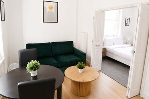 Ein Zimmer mit einem grünen Sofa, einem runden Holztisch mit einer Pflanze, einem weißen Bett und einer Tür zu einem anderen Raum.
