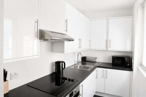 A modern kitchen with white cabinets, a black countertop, a stainless steel range hood, a kettle, a sink, and a microwave. - Bright 1BR in Fitzrovia, Steps from Oxford Street (London)