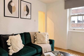 A room with a green sofa, two framed abstract art pieces, a window with a view of buildings, and a door leading to another room. - Elegant 1BR, Fitzrovia, Steps from West End (London)