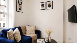Een woonkamer met een blauwe bank, een bijzettafeltje met tijdschriften, een vaas met gedroogde planten en een aan de muur bevestigde televisie.
