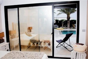 A bedroom with a bed, bedside table, lamp, and a view of a swimming pool through a glass door.