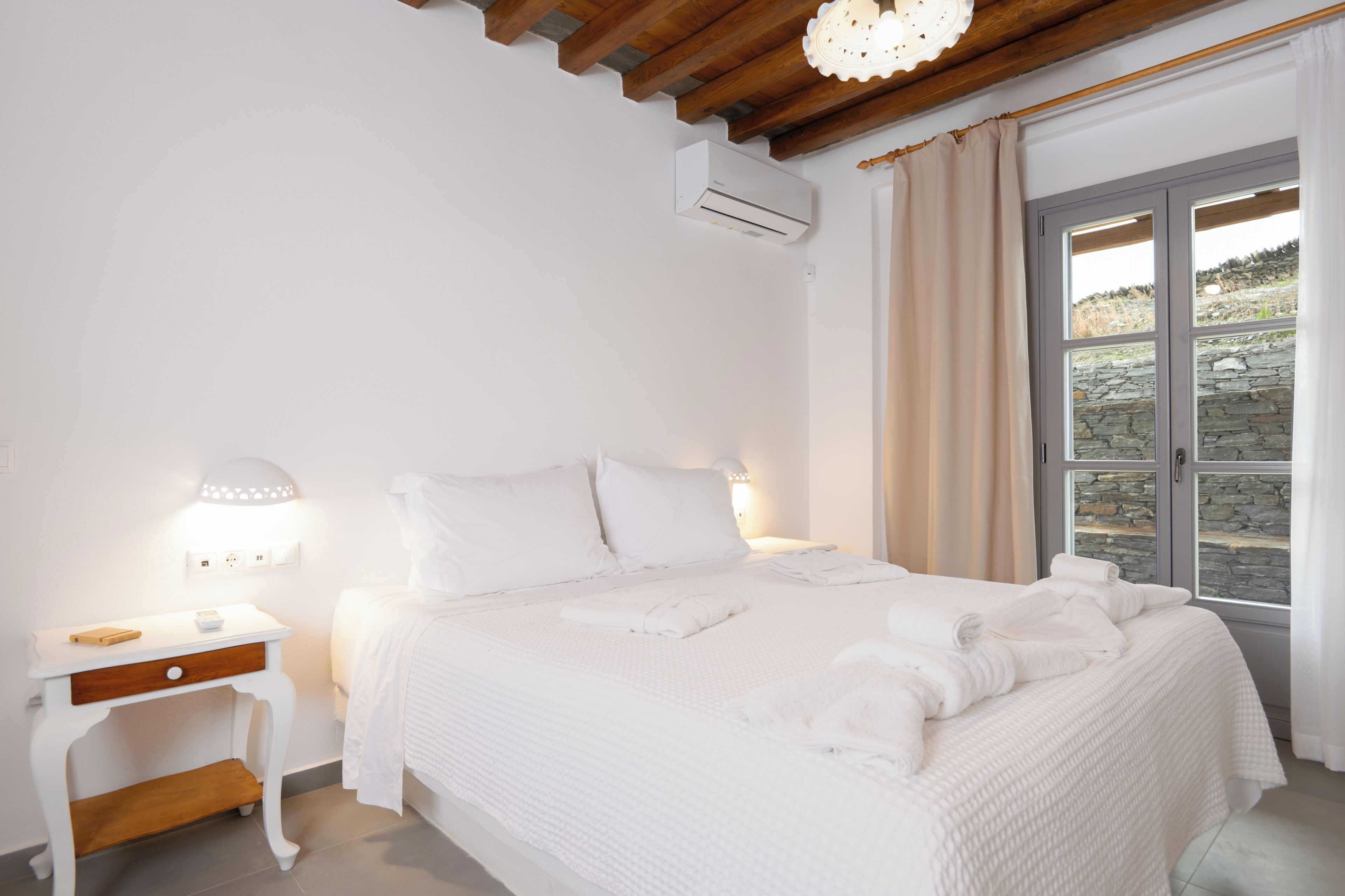 A white bedroom with a bed, bedside table, lamp, and a window with curtains.