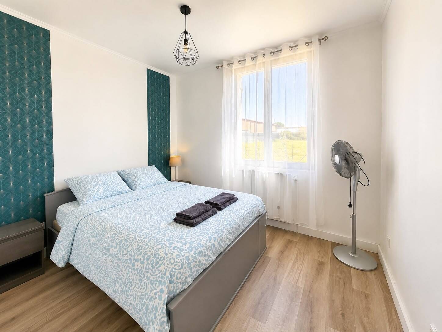 A bedroom with a bed, a standing fan, a window with curtains, and a wall with a geometric pattern.