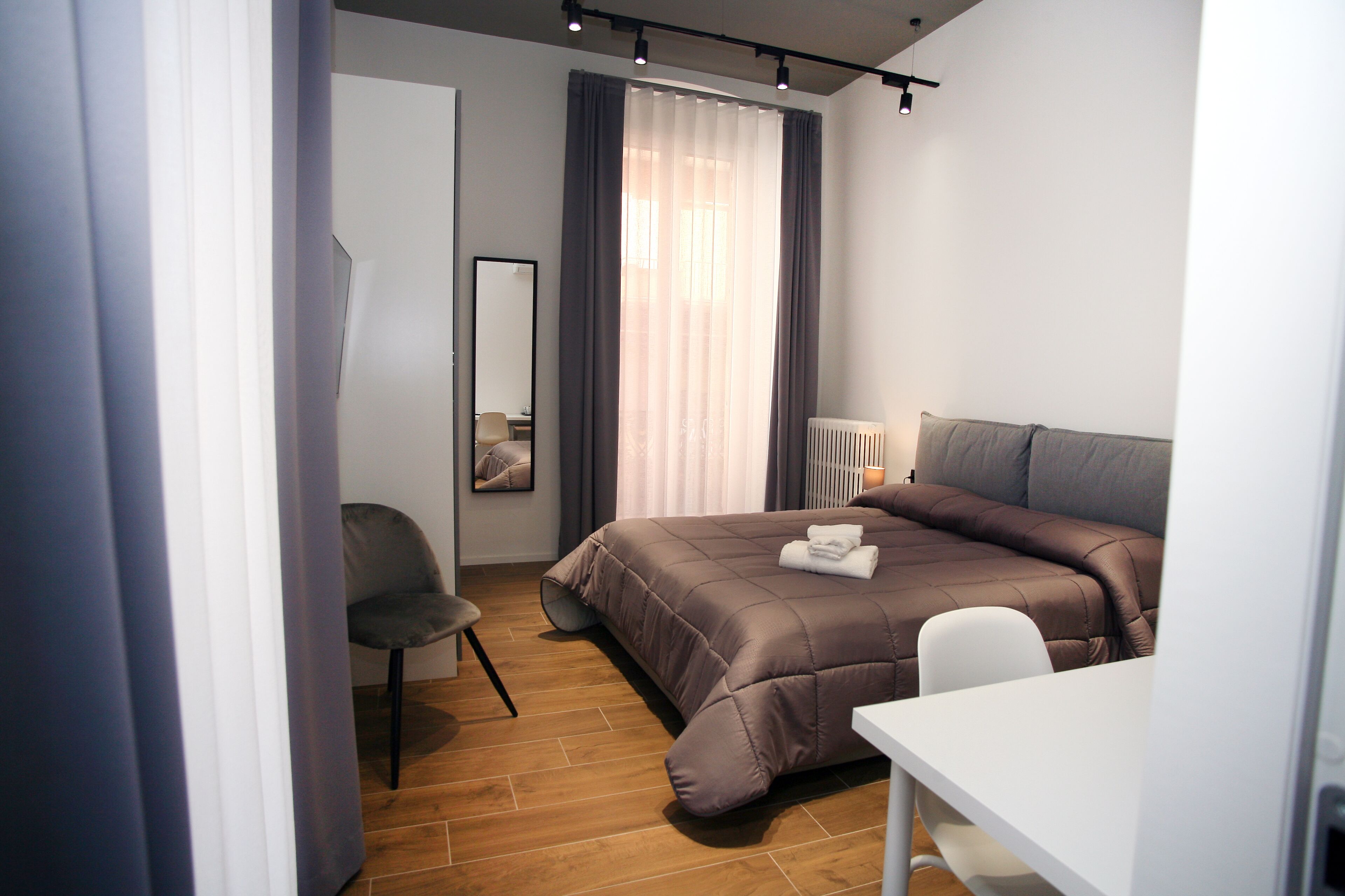 A modern bedroom with a bed, a desk, a chair, a mirror, and a window with curtains.