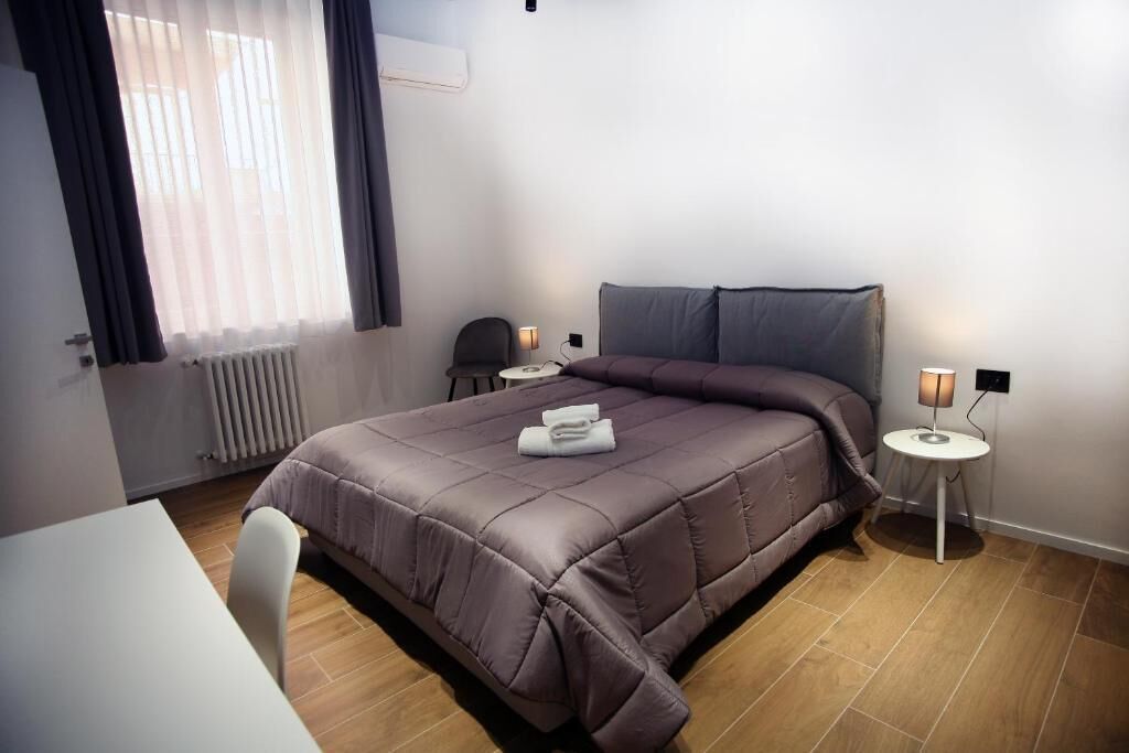 Un dormitorio con cama, una silla, una mesa, una lámpara y un ventanal con cortinas.