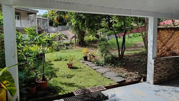 A covered outdoor area with a concrete floor, potted plants, a small grassy patch, and a stone pathway. - GROUND-FLOOR HOUSE—GARDEN—ROOMS FOR RENT AND A TERRACE WITH A PING-PONG TABLE (Rivière-Salée)