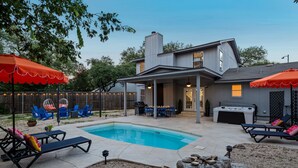 A backyard with a swimming pool, lounge chairs, a hot tub, and a covered patio area.