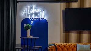 A room with a neon sign reading 'Aloha Ya'll,' a small table with stools, a tufted orange sofa, and a large flat-screen TV mounted on the wall.
