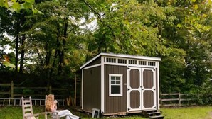 A small wooden shed with a window and a door, a rocking chair, and a fire pit in a grassy area.