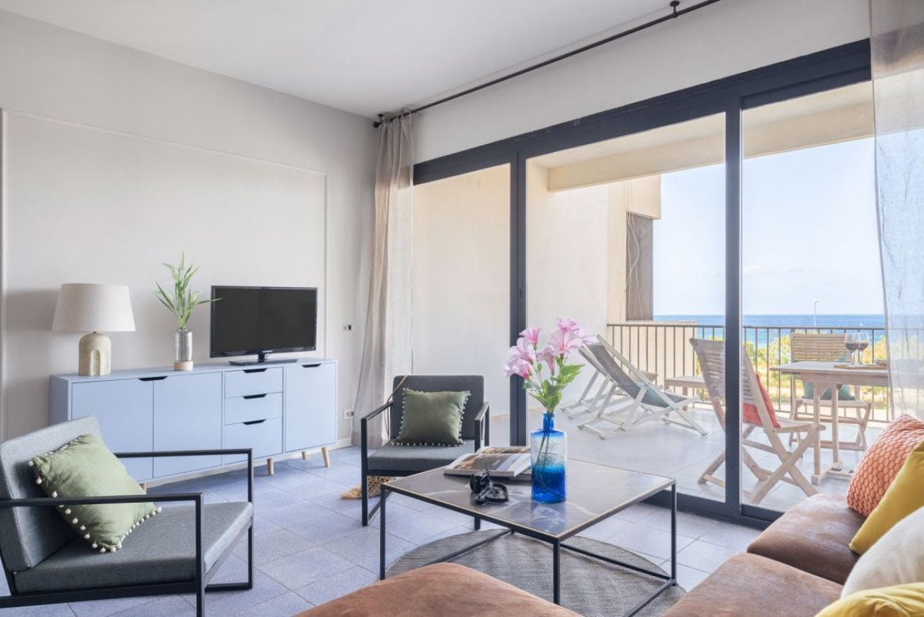 Una sala de estar moderna con sofá, sillón, mesa de centro, televisor y un balcón con mobiliario exterior.