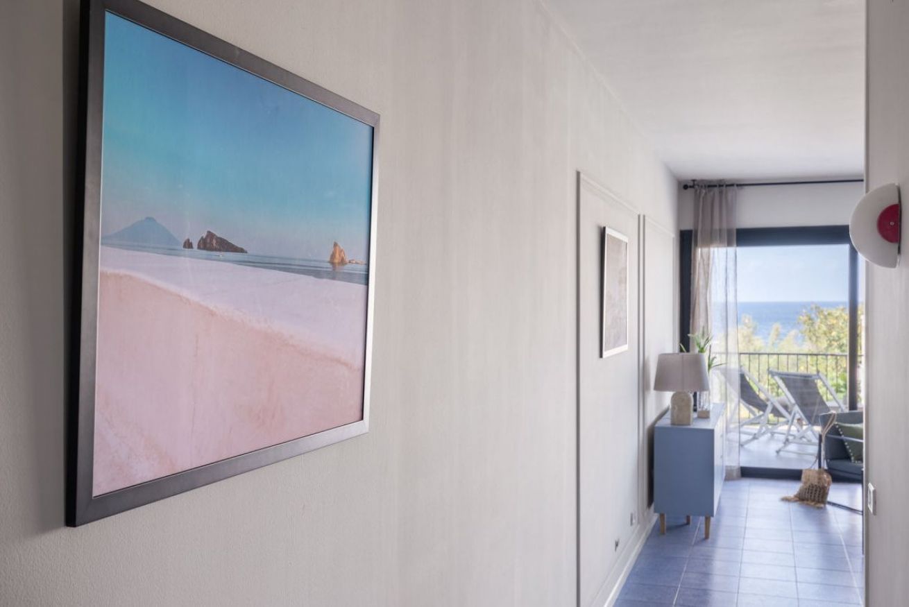 A hotel corridor with a large framed picture of a beach, a small side table with a lamp, and a balcony with chairs.