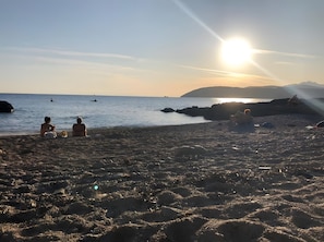 Ein Strand mit Menschen, die am Sand sitzen, einem Sonnenuntergang im Hintergrund und felsigem Gelände.
