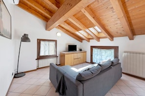 Una stanza con soffitto in legno alto, un divano, una scrivania con un computer e un termosifone.