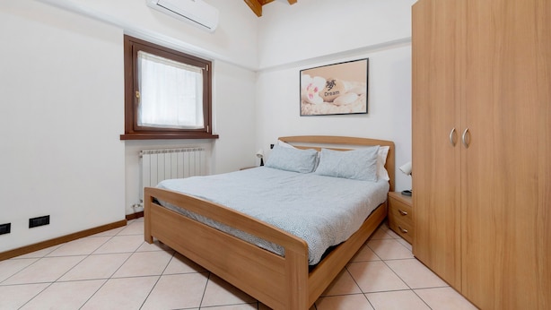 A bedroom with a wooden bed, a window, an air conditioner, a radiator, a wardrobe, and a framed picture on the wall. - Apartment 'La Mansarda Sul Lago' with Lake View, Wi-Fi and Air Conditioning (Lovere)