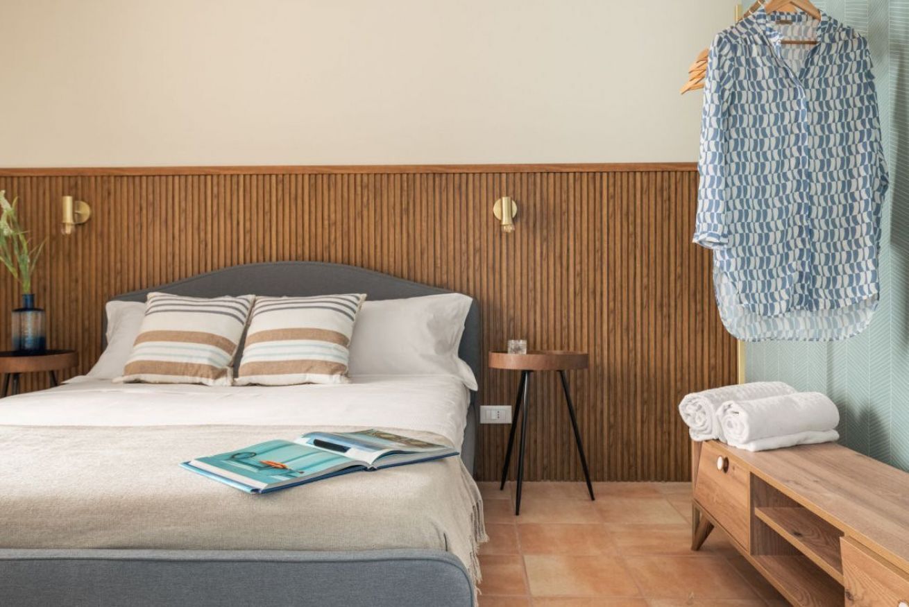 A neatly arranged bedroom with a bed, pillows, a bedside table, a small stool, and a bench with towels.