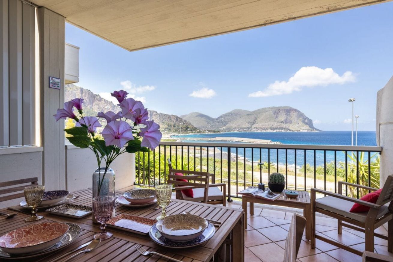 A dining area with a table set for two, a vase with flowers, and a view of the ocean and mountains.