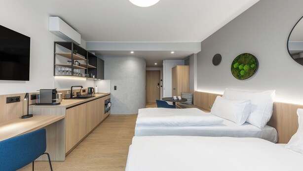 A modern hotel room with a kitchenette, two beds, a dining area, and a large mirror.