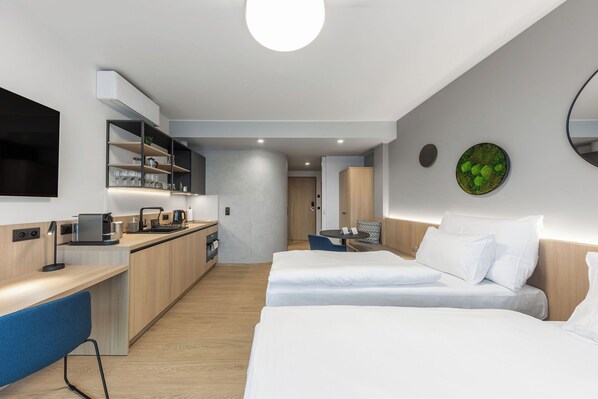A modern hotel room with a kitchenette, two beds, a dining area, and a large mirror.