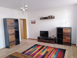 A modern living room with a flat-screen TV, wooden cabinets, a colorful rug, and a hanging light fixture.
