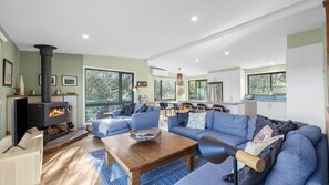 A modern living room with a fireplace, blue sofas, a wooden coffee table, and a kitchen in the background. - Awel -Y- Mor - Pet friendly beach retreat (Moggs Creek)