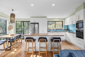 A modern kitchen with a central island, bar stools, a dining area with a table and chairs, and large windows offering a view of greenery. - Awel -Y- Mor (Moggs Creek)