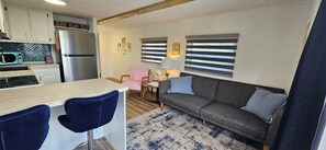 Ein kompakter Wohnraum mit Küchenzeile, einer Couch, einem Sessel und einem kleinen Tisch mit Lampe.
