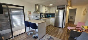 Eine moderne Küche mit einem Edelstahl-Kühlschrank, einer Mikrowelle und einem Gasherd. Zwei blaue Barhocker und ein Holztisch sind ebenfalls vorhanden.