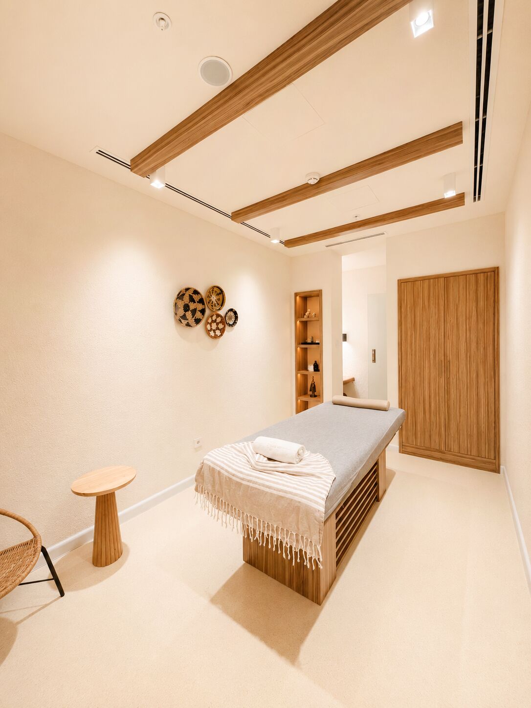 A modern spa room with a massage table, wooden beams, a small round table, and a woven chair.